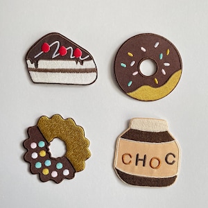 Könnte beinhalten: Vier gestickte Aufnäher mit Dessertmotiven. Enthält ein Stück Schokoladenkuchen mit roten Kirschen, einen Schokoladen-Donut mit Streuseln, einen Keks mit gewelltem Rand und ein Glas Schokoladenaufstrich mit dem Wort "CHOC".
