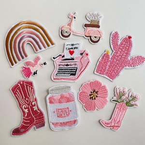 Może przedstawiać: Kolekcja haftowanych naszywek w odcieniach różu, czerwieni i brązu. Wzory obejmują tęczę, ptaka, skuter, maszynę do pisania z napisem "I love you", kaktus, kowbojski but, słoik z napisem "love", kwiat i but z kwiatami.