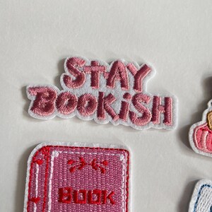 May include: Embroidered patches with a book theme. The top patch reads "STAY BOOKISH" in pink lettering with a white border. Other patches include a stack of books and a book with the word "BOOK" on it.