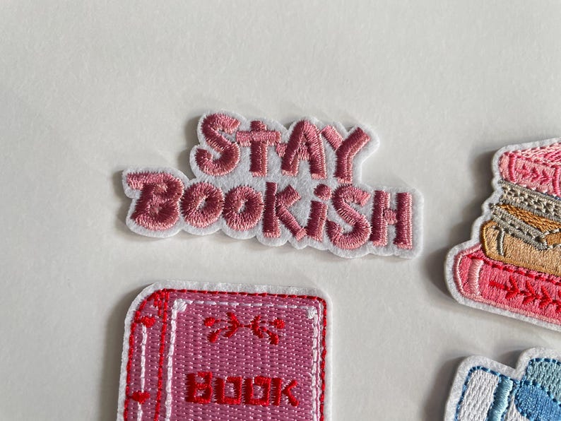 May include: Embroidered patches with a book theme. The top patch reads "STAY BOOKISH" in pink lettering with a white border. Other patches include a stack of books and a book with the word "BOOK" on it.