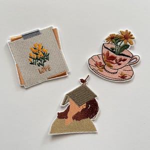Op de afbeelding: Drie geborduurde patches op een witte achtergrond. Eén patch heeft een bloemenontwerp met het woord "LOVE". Een andere toont een theekopje met bloemen. De derde patch toont een persoon met een hoed.