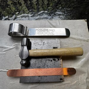 Puede incluir: Un conjunto de herramientas met&aacute;licas, que incluye una llave inglesa plateada con el texto "ZOOM PRECISION", un martillo peque&ntilde;o con mango de madera y una tira de cobre. Las herramientas est&aacute;n dispuestas sobre una superficie gris oscuro.