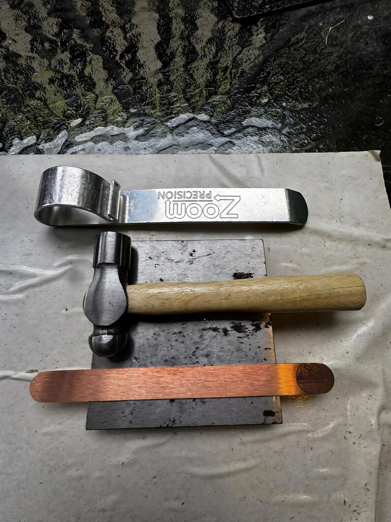 Puede incluir: Un conjunto de herramientas met&aacute;licas, que incluye una llave inglesa plateada con el texto "ZOOM PRECISION", un martillo peque&ntilde;o con mango de madera y una tira de cobre. Las herramientas est&aacute;n dispuestas sobre una superficie gris oscuro.