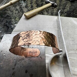 Puede incluir: Un brazalete de cobre martillado con una superficie texturizada. El brazalete abierto est&aacute; sobre una superficie met&aacute;lica, con martillos y herramientas en el fondo. Un dise&ntilde;o estampado es visible cerca de la abertura.