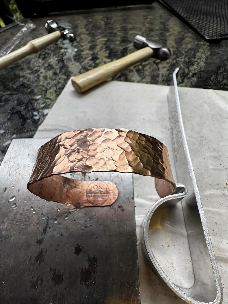 Puede incluir: Un brazalete de cobre martillado con una superficie texturizada. El brazalete abierto est&aacute; sobre una superficie met&aacute;lica, con martillos y herramientas en el fondo. Un dise&ntilde;o estampado es visible cerca de la abertura.