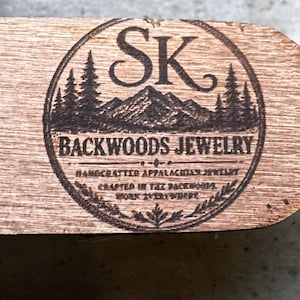 Puede incluir: Placa de madera con un logotipo circular con las letras "SK" sobre una cordillera y pinos. Las palabras "BACKWOODS JEWELRY" est&aacute;n sobre el texto "HANDCRAFTED APPALACHIAN JEWELRY CRAFTED IN THE BACKWOODS WORK EVERYWHERE."