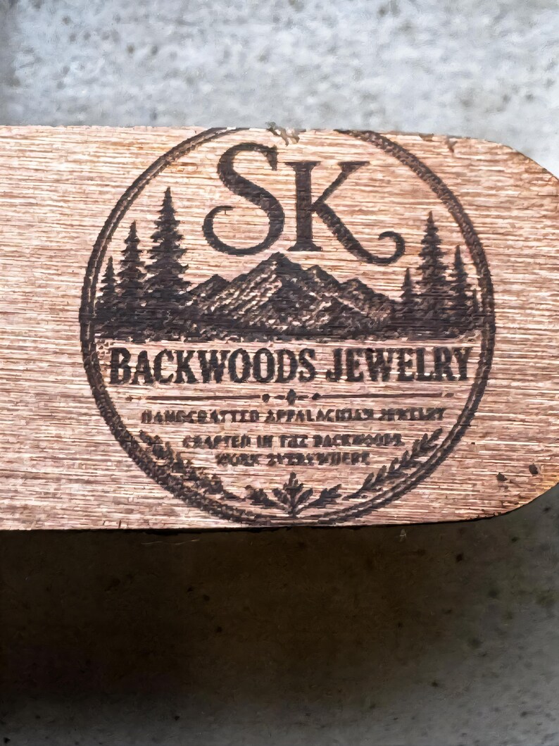 Puede incluir: Placa de madera con un logotipo circular con las letras "SK" sobre una cordillera y pinos. Las palabras "BACKWOODS JEWELRY" est&aacute;n sobre el texto "HANDCRAFTED APPALACHIAN JEWELRY CRAFTED IN THE BACKWOODS WORK EVERYWHERE."
