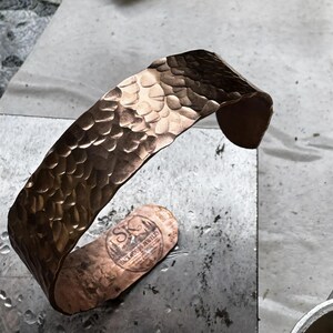 Puede incluir: Un brazalete de cobre martillado con una superficie texturizada. El brazalete tiene forma curva y est&aacute; estampado con "Backwoods Jewelry". El brazalete mide aproximadamente 3,8 cm de ancho.