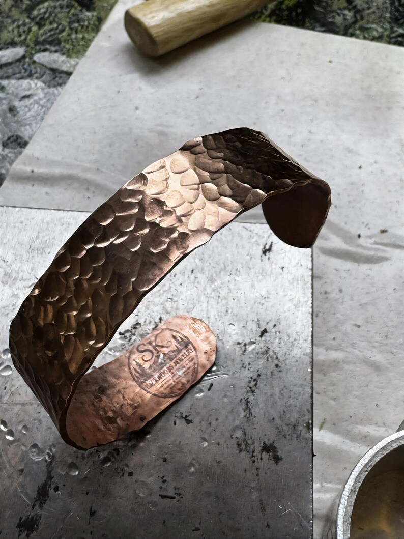 Puede incluir: Un brazalete de cobre martillado con una superficie texturizada. El brazalete tiene forma curva y est&aacute; estampado con "Backwoods Jewelry". El brazalete mide aproximadamente 3,8 cm de ancho.