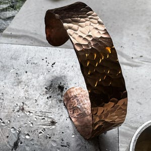 Puede incluir: Una pulsera de pu&ntilde;o de cobre martillado con una superficie texturizada. El dise&ntilde;o de pu&ntilde;o abierto presenta una serie de peque&ntilde;as indentaciones irregulares en su superficie. La pulsera tiene un brillo met&aacute;lico c&aacute;lido, que refleja la luz. El pu&ntilde;o mide aproximadamente 5 cm de ancho.