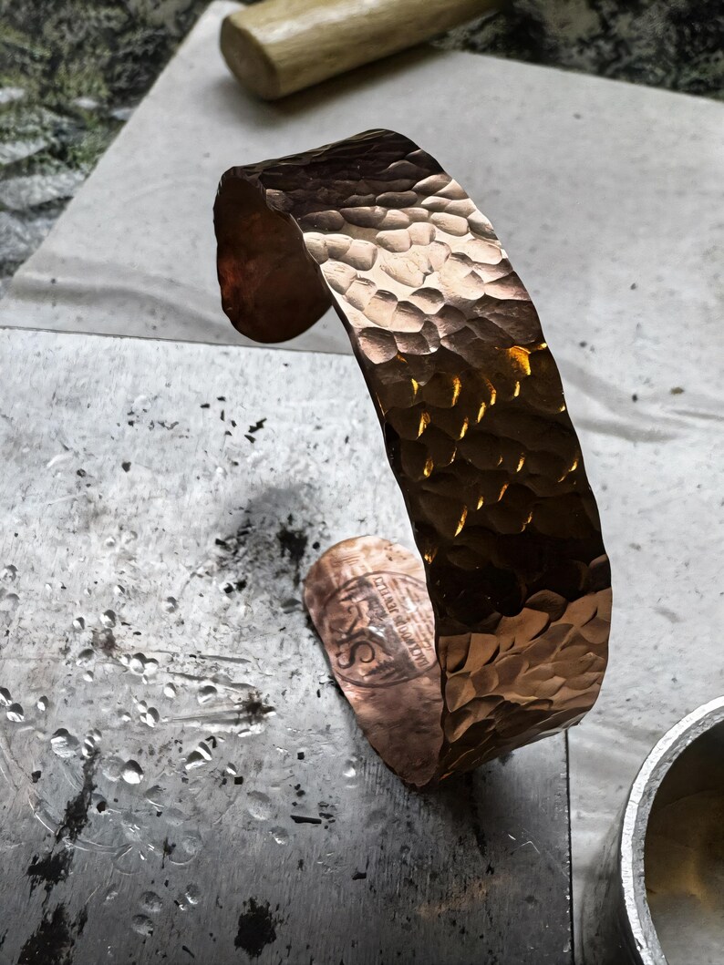Puede incluir: Una pulsera de pu&ntilde;o de cobre martillado con una superficie texturizada. El dise&ntilde;o de pu&ntilde;o abierto presenta una serie de peque&ntilde;as indentaciones irregulares en su superficie. La pulsera tiene un brillo met&aacute;lico c&aacute;lido, que refleja la luz. El pu&ntilde;o mide aproximadamente 5 cm de ancho.