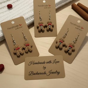 May include: Gingerbread man earrings on display. Each pair of earrings is attached to a brown card with the text "Handmade with love." The cards are arranged on a white surface with red berries and cinnamon sticks.