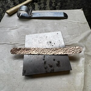 Puede incluir: Una barra de metal de cobre con una superficie texturizada descansa sobre un yunque de metal. Una herramienta de metal plateado y un martillo con mango de madera est&aacute;n en el fondo. Las herramientas sugieren trabajos en metal o joyer&iacute;a.