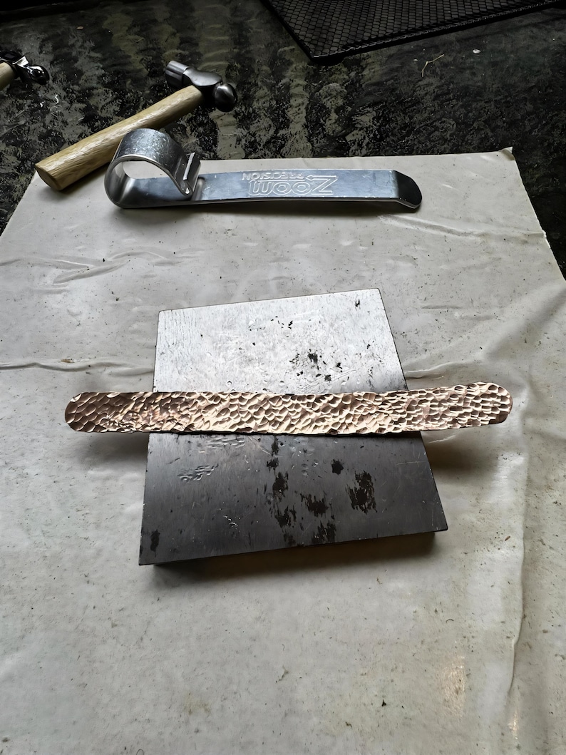 Puede incluir: Una barra de metal de cobre con una superficie texturizada descansa sobre un yunque de metal. Una herramienta de metal plateado y un martillo con mango de madera est&aacute;n en el fondo. Las herramientas sugieren trabajos en metal o joyer&iacute;a.