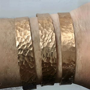 May include: Four hammered gold-tone metal bracelets are worn on a wrist. The bracelets have a textured surface and a flat, rectangular shape. The bracelets are different shades of gold, ranging from light to dark.