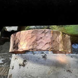Puede incluir: Un brazalete de cobre martillado. El brazalete ancho tiene una superficie texturizada con un patr&oacute;n martillado. El cobre tiene un tono marr&oacute;n rojizo c&aacute;lido con &aacute;reas de tonos dorados m&aacute;s claros. El brazalete se muestra sobre una superficie met&aacute;lica.