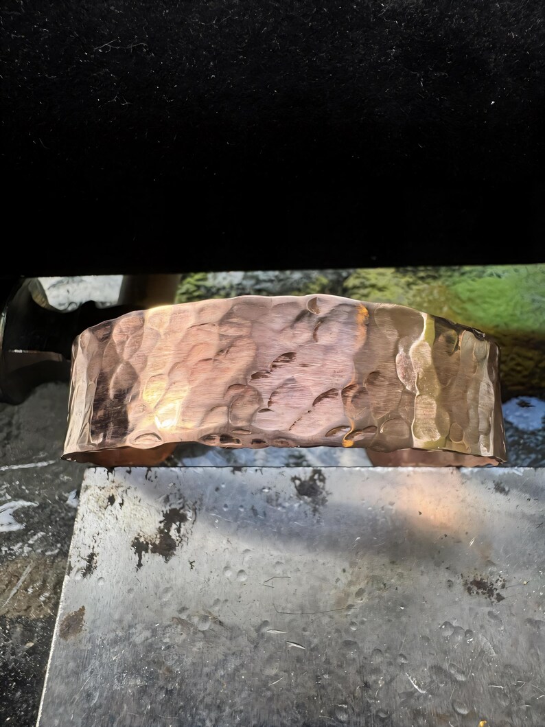 Puede incluir: Un brazalete de cobre martillado. El brazalete ancho tiene una superficie texturizada con un patr&oacute;n martillado. El cobre tiene un tono marr&oacute;n rojizo c&aacute;lido con &aacute;reas de tonos dorados m&aacute;s claros. El brazalete se muestra sobre una superficie met&aacute;lica.