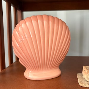 May include: A peach-colored ceramic seashell sculpture. The sculpture has a ribbed texture and a rounded base. The seashell is displayed on a wooden shelf. The sculpture is a decorative item.
