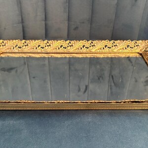 May include: A rectangular gold-toned vanity tray with a mirrored surface. The tray features ornate, detailed scrollwork around the edges. The tray is set on a dark blue surface.