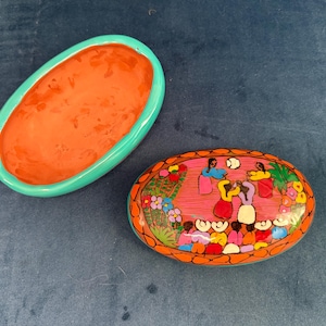 May include: Oval-shaped, hand-painted trinket box with a lid featuring a colorful scene of people dancing. The box has a turquoise rim and a bright orange interior. The lid is decorated with flowers and figures.