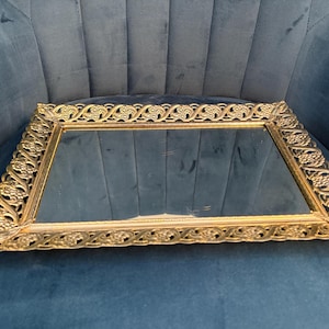 May include: Rectangular gold-toned vanity tray with a mirrored surface. The tray features an ornate, repeating floral design around the edges. The tray is resting on a blue velvet surface.