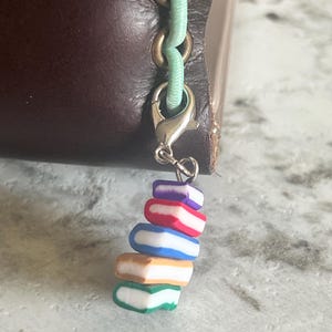 May include: A stack of colorful miniature books charm attached to a silver clasp and a light green cord. The books have white pages and covers in red, blue, orange, purple, and green. The charm is attached to a brown leather item.