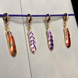 May include: Four gold-toned metal feather charms with lobster claw clasps, each featuring distinct enamel patterns: one with orange and black scales, one with purple and white stripes, one with purple and black stripes, and one with red and black.