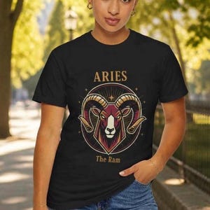 May include: Black t-shirt featuring a detailed illustration of a ram's head within a circular design. The word "ARIES" is arched above the ram, with "The Ram" below. The design is in shades of burgundy, gold, and white.