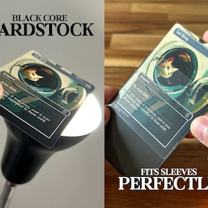 May include: A black core cardstock card is held up to a light, showcasing the card's design. The card features an illustration with a cat and the text "Sol Ring" and "Artifact". The text "BLACK CORE CARDSTOCK" and "FITS SLEEVES PERFECTLY" are also visible.
