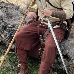 May include: A medieval-style outfit featuring a cream-colored tunic, brown pants, and leather boots. The ensemble includes a sword, leather arm guards, and a walking stick. The outfit is suitable for cosplay or historical reenactment.