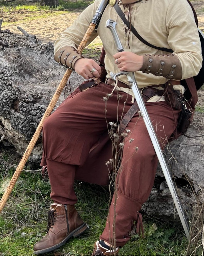 May include: A medieval-style outfit featuring a cream-colored tunic, brown pants, and leather boots. The ensemble includes a sword, leather arm guards, and a walking stick. The outfit is suitable for cosplay or historical reenactment.