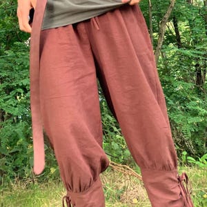 May include: Rust-colored, loose-fitting pants with gathered ankles and drawstring ties. The pants are made of a lightweight fabric and have a relaxed, flowing silhouette. The pants are part of a medieval or renaissance-inspired outfit.