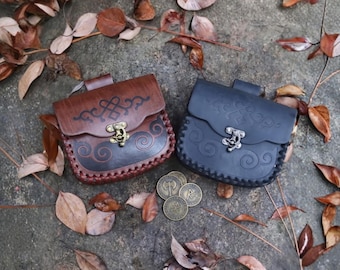 May include: Two leather pouches, one brown and one dark blue, with decorative clasps and embossed designs. The pouches are accompanied by several bronze coins. The scene is set on a stone surface with scattered leaves.