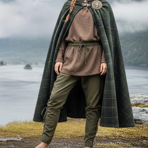 May include: A green plaid cloak with a decorative clasp, worn over a brown tunic and olive green pants. The outfit has a medieval or fantasy theme, with a focus on historical or cosplay attire.