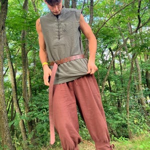 May include: A sleeveless olive green tunic with a lace-up neckline and a brown leather belt. The outfit includes rust-colored, loose-fitting pants with ankle ties. The clothing is suitable for historical reenactment or costume events.