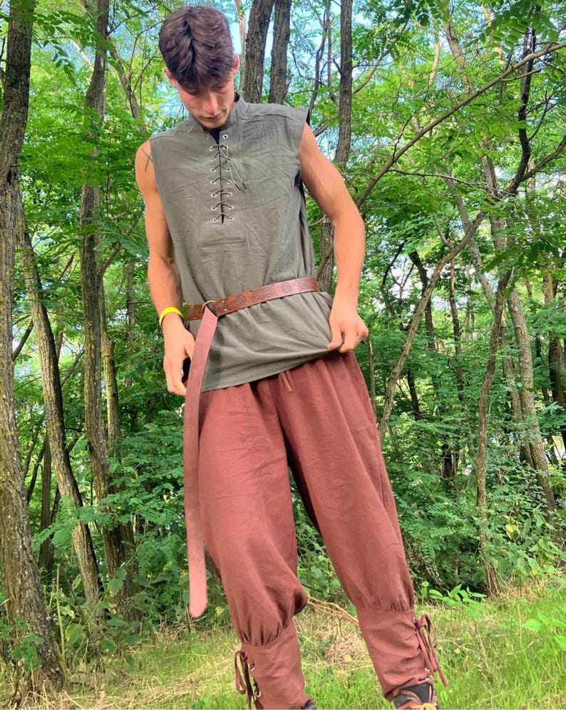 May include: A sleeveless olive green tunic with a lace-up neckline and a brown leather belt. The outfit includes rust-colored, loose-fitting pants with ankle ties. The clothing is suitable for historical reenactment or costume events.