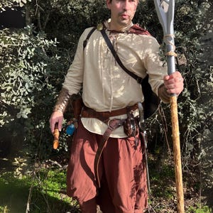Peut inclure: Une personne en costume fantastique, v&ecirc;tue d'une chemise cr&egrave;me &agrave; manches longues, d'un pantalon marron et d'accessoires en cuir. Elle tient un b&acirc;ton avec un sommet argent&eacute; et une base en bois. Le costume comprend une ceinture marron, un sac &agrave; bandouli&egrave;re et des prot&egrave;ge-bras.