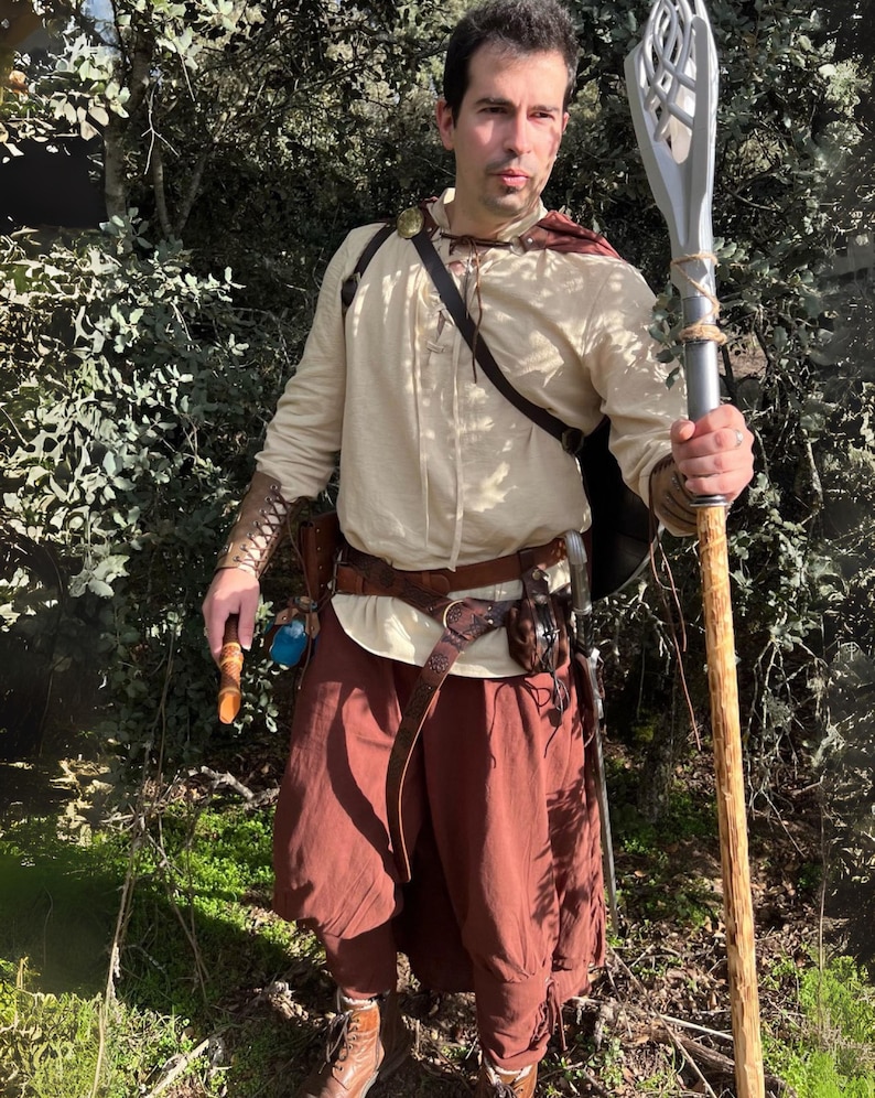 Peut inclure: Une personne en costume fantastique, v&ecirc;tue d'une chemise cr&egrave;me &agrave; manches longues, d'un pantalon marron et d'accessoires en cuir. Elle tient un b&acirc;ton avec un sommet argent&eacute; et une base en bois. Le costume comprend une ceinture marron, un sac &agrave; bandouli&egrave;re et des prot&egrave;ge-bras.