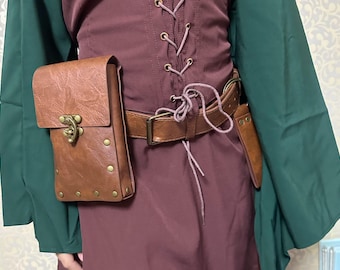 May include: A brown leather pouch with a flap and brass-colored hardware is attached to a brown belt. The belt is worn over a burgundy top with a lace-up front. A smaller brown leather sheath is also attached to the belt.