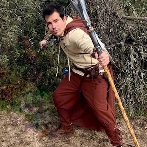 Peut inclure: Une personne en costume fantastique, tenant une lance et une petite lame. La tenue comprend une chemise beige, un pantalon marron, une cape marron et des bottes marron. La lance a une lame argent&eacute;e et un manche en bois.