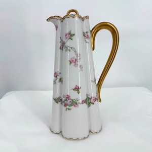 May include: White porcelain pitcher with a gold handle and rim. The pitcher is decorated with pink roses and green leaves. The handle and rim are gold. The pitcher is approximately 10 inches tall.