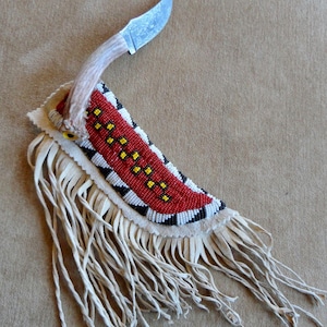 May include: A handcrafted knife with a decorative beaded sheath. The sheath features red, black, white, and yellow beadwork, with a fringe of cream-coloured leather. The knife has a silver blade and a light-coloured antler handle.