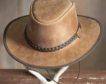 May include: A brown leather hat with a wide brim and a braided band. The hat has a dark brown trim and a chin strap. The hat is displayed on a white antler against a rustic wooden background. The hat is a classic western style.