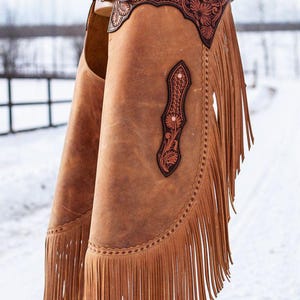 Op de afbeelding: Bruine leren chaps met decoratieve bloemmotieven en franjes. De chaps hebben een gespsluiting en worden gepresenteerd tegen een besneeuwde achtergrond. Het leer heeft een verweerde uitstraling.