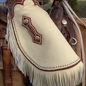 Op de afbeelding: Cr&egrave;me leren chaps met bruine sierlijsten en franjes. De chaps hebben een bloemmotief en zilverkleurige versieringen. Deze chaps zijn ontworpen om op te paardrijden. Western-thema product.