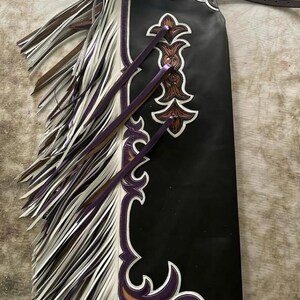 Op de afbeelding: Zwarte leren chaps met decoratieve bruine en witte bloemmotieven. De chaps hebben witte en paarse franjes en paarse accenten. Een bruine leren riem is aan de bovenkant bevestigd.