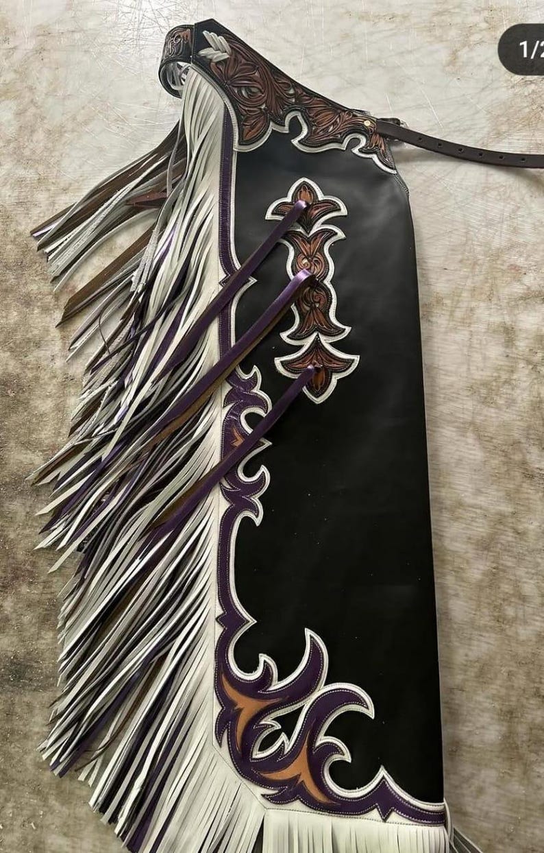 Op de afbeelding: Zwarte leren chaps met decoratieve bruine en witte bloemmotieven. De chaps hebben witte en paarse franjes en paarse accenten. Een bruine leren riem is aan de bovenkant bevestigd.