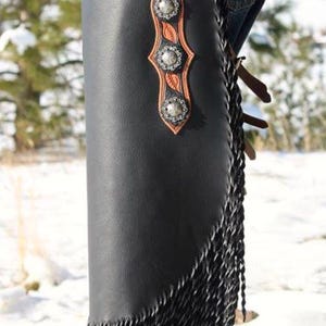 Op de afbeelding: Zwarte leren chaps met zilveren studs en een bruin bloemmotief. De chaps hebben een franje langs de zijkant en worden over een spijkerbroek gedragen. Een western item.