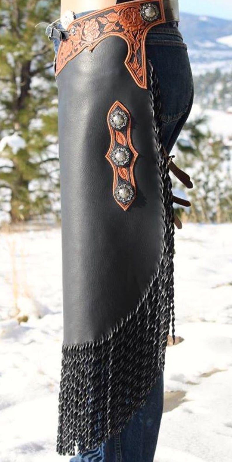 Op de afbeelding: Zwarte leren chaps met zilveren studs en een bruin bloemmotief. De chaps hebben een franje langs de zijkant en worden over een spijkerbroek gedragen. Een western item.