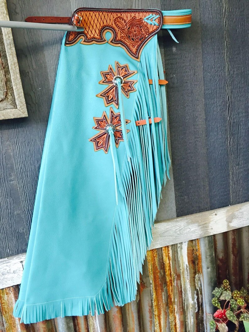 Op de afbeelding: Turquoise leren chaps met decoratieve bloemen- en sterontwerpen. De chaps hebben lange franjes langs de zijkanten en onderkant, met bruine leren accenten en een bewerkte leren juk. De chaps hangen aan een houten constructie.